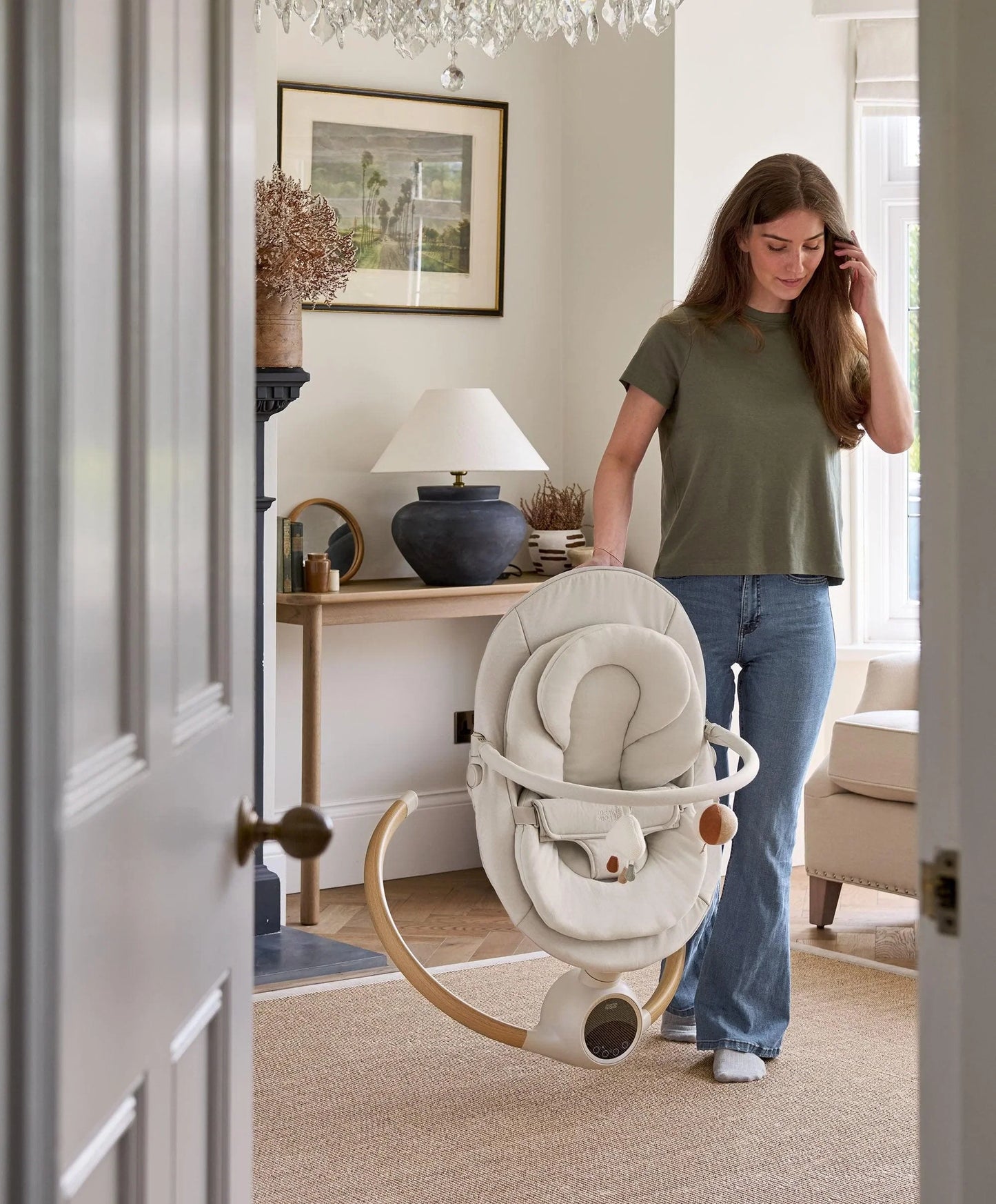 Cream baby swing with plush seat and hanging toy arch, upright near doorway.