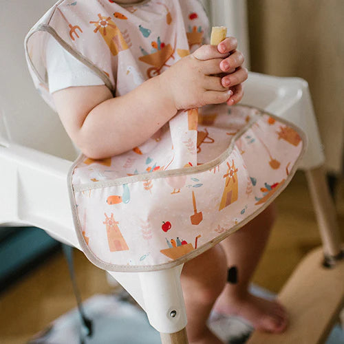 Child wearing light pink farmer apron with farm icons, bib flap at high chair