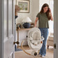 Cream baby swing with plush seat and hanging toy arch, upright near doorway.