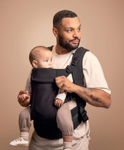 Black baby carrier with front panel, worn by parent holding seated infant