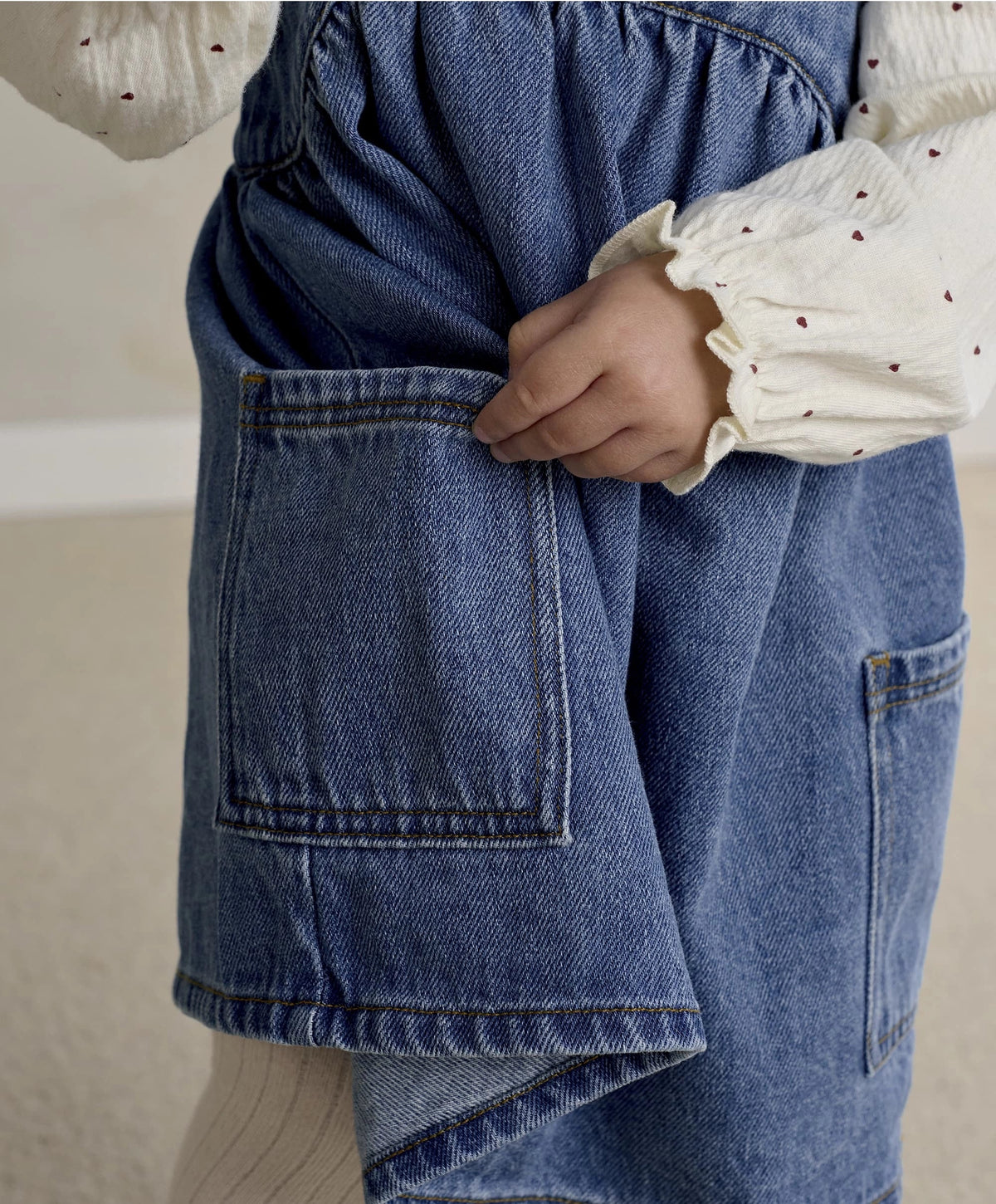 Blue denim pinafore dress with front patch pockets and topstitching over white bodysuit
