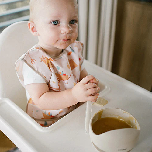 Child wearing light pink farmer apron with colorful farm icons at high chair