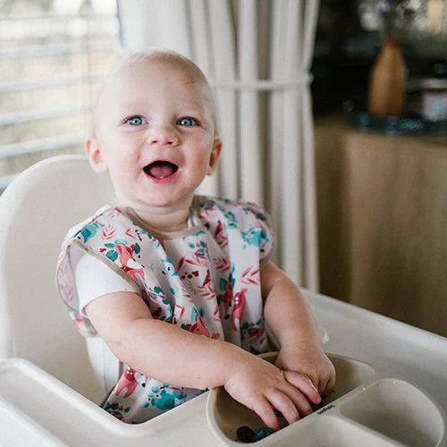Kids sleeveless apron with forest animal pattern in pink and teal, worn in high chair