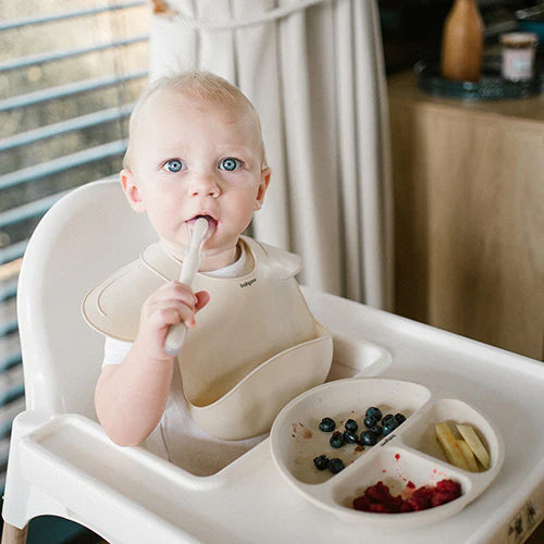 Beige silicone bib worn by baby, with wide curved front and neck strap